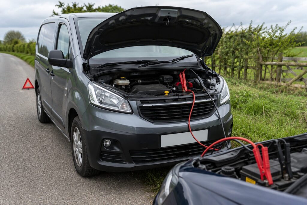 How to Jump Start A Car 1 Grey panel van parked on a rural roadside with its bonnet open, connected to another vehicle by jump leads for a battery boost. A warning triangle is placed behind the van on the road, with hedgerows and fields visible in the countryside background under a cloudy sky.