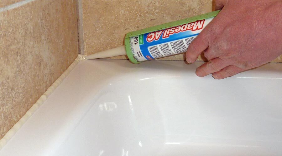 A person applies a bead of white silicone sealant with a caulking gun along the edge of a bathtub where it meets a tiled wall.