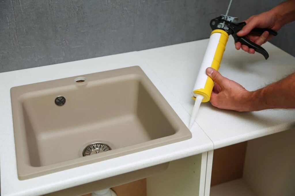 A person applies sealant with a caulking gun along the joint of a white countertop during the installation of an inset kitchen sink.