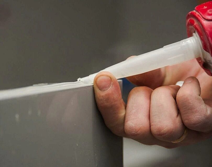 Close-up of a person applying a bead of white sealant along the edge of a panel using a cartridge gun, with the nozzle pressed against the joint for a neat finish.