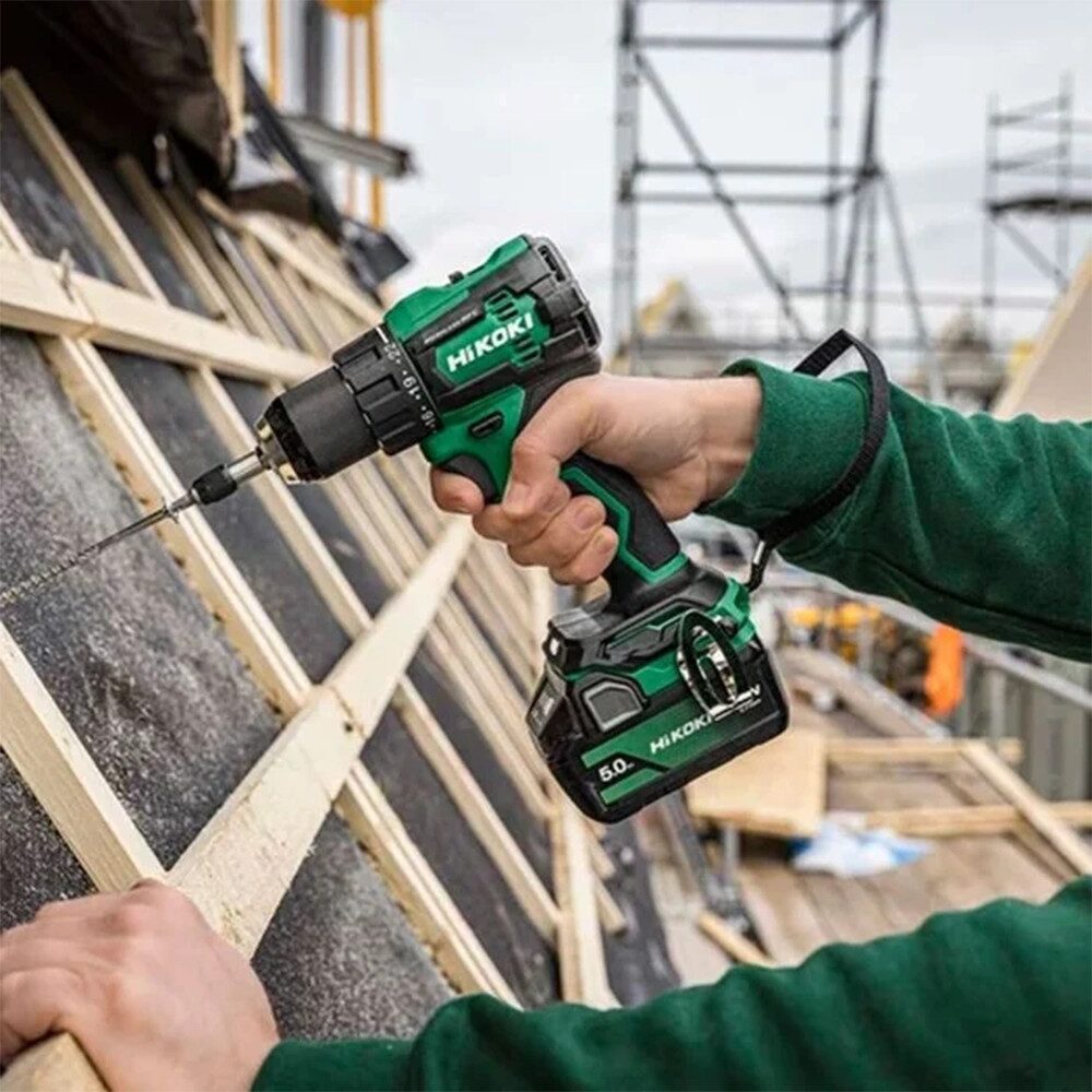 A green HiKOKI cordless combi drill is being used to drive a fixing into timber roof battens on a construction site. The drill is held in one hand and fitted with a driver bit, while the other hand steadies the timber. Scaffolding and building materials are visible in the blurred background, indicating an active roofing or framing project. The image demonstrates the drill being used for fastening and general construction work.