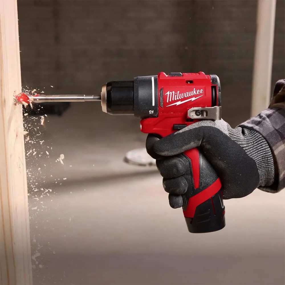 Close up of a compact red Milwaukee cordless drill being used to drill into a wooden stud. A gloved hand grips the tool while a drill bit cuts into the timber, creating visible wood shavings and dust. The background is blurred, suggesting an indoor construction or workshop environment. The image demonstrates the drill being used for light duty drilling in wood.