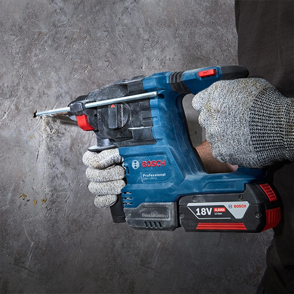 A blue Bosch Professional cordless SDS Plus rotary hammer drill is being used to drill into a concrete wall. The tool is held with both hands while an SDS drill bit bores into the masonry surface, creating visible dust around the hole. The drill features a side handle for improved control and an 18V battery pack. The image demonstrates heavy duty drilling in concrete and masonry applications using hammer action.