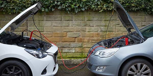 How to Jump Start A Car 14 Two cars parked facing each other with their bonnets open, connected by red and black jump leads between the engine bays. The vehicles are positioned in front of a stone wall, illustrating a battery jump start procedure using a donor vehicle.