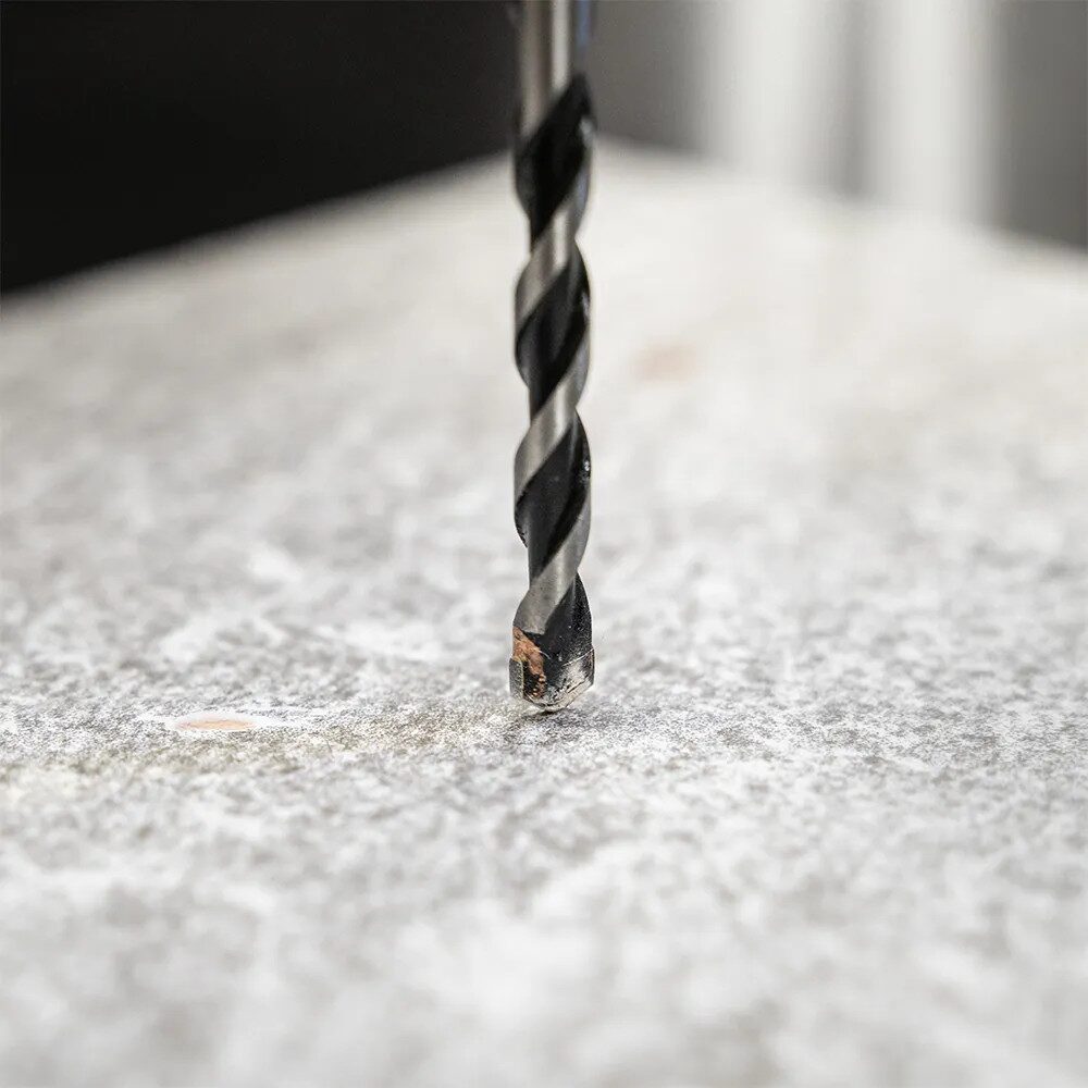 Drill Bits Buying Guide: Choosing the Right Drill Bit for Every Material 3 Close up photograph of a carbide tipped masonry drill bit positioned vertically against a light grey concrete or stone surface. The tungsten carbide cutting tip is visible at the end of the spiral fluted bit, showing the distinctive chisel shaped head used for drilling into masonry materials. The background is softly blurred, drawing attention to the drill bit tip and its contact point with the surface.