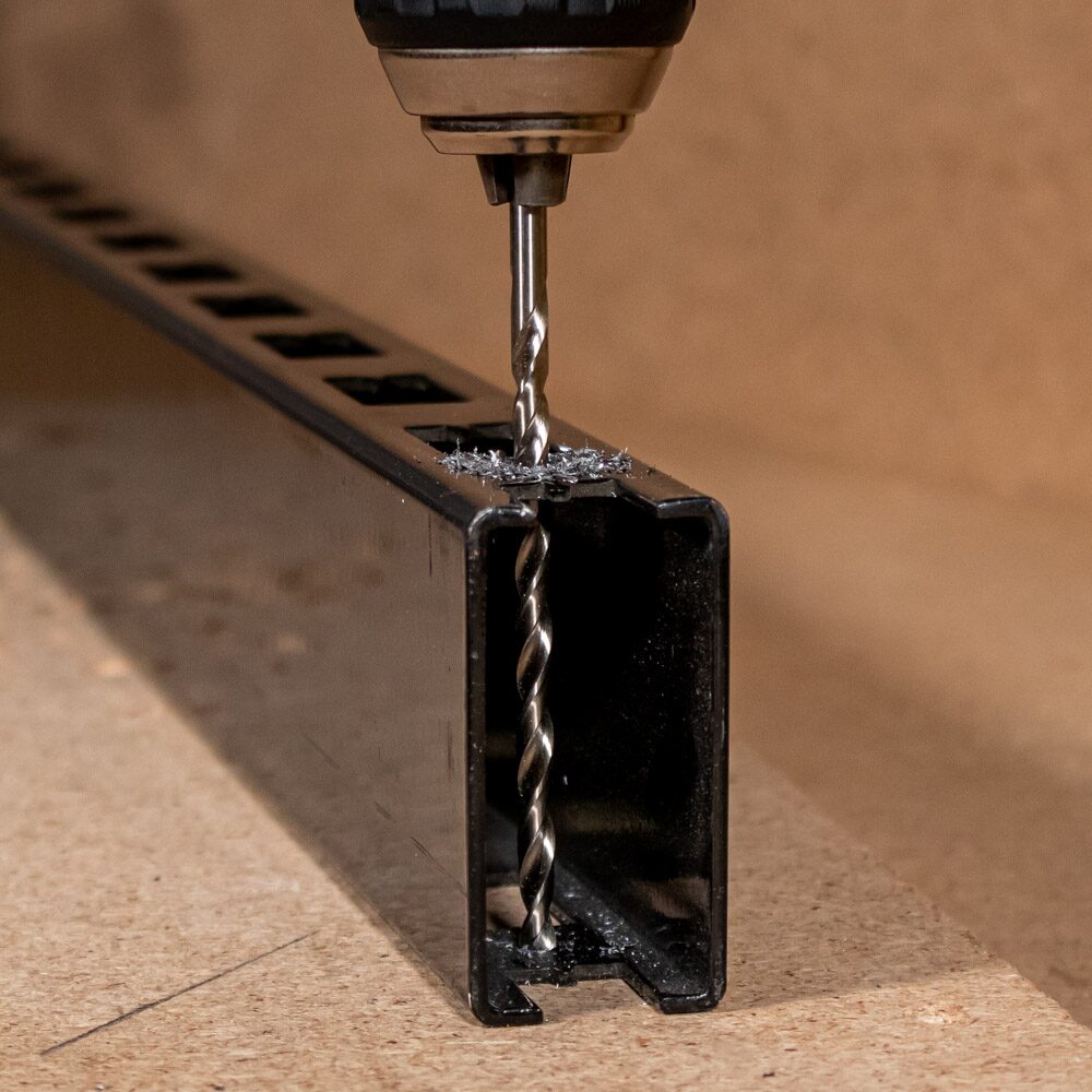 Drill Bits Buying Guide: Choosing the Right Drill Bit for Every Material 5 Close up image of a metal drill bit drilling through a black steel box section. The bit is mounted in a power drill and is cutting through the top surface of the hollow metal profile, producing metal swarf around the hole. The open end of the box section reveals the drill bit passing through the material, demonstrating metal drilling in a fabrication or workshop setting.