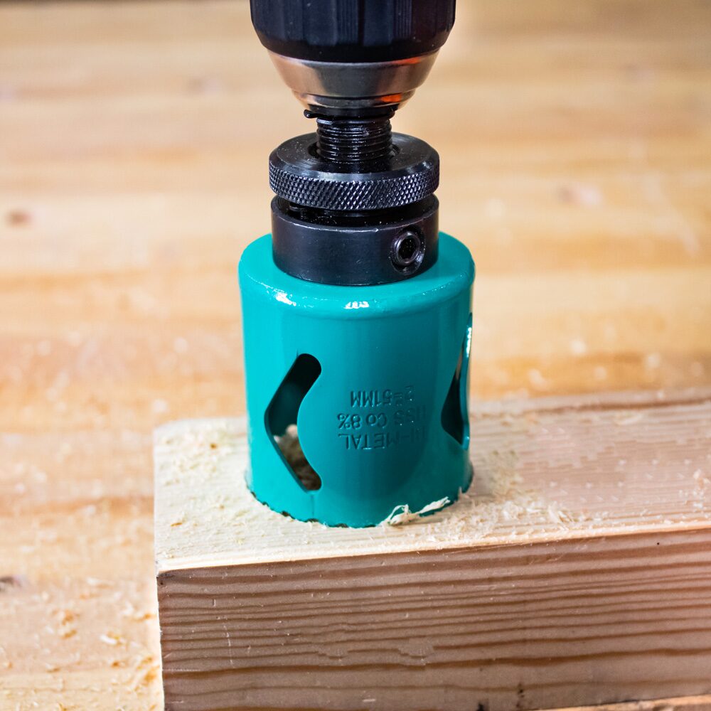 Drill Bits Buying Guide: Choosing the Right Drill Bit for Every Material 18 Close up image of a hole saw attached to a power drill cutting a large circular hole into a piece of timber. The cylindrical hole saw features side slots for chip clearance and is fitted with a central pilot drill bit to guide the cut. Fine sawdust surrounds the cutting area as the hole saw bores into the wood, demonstrating its use for creating large diameter holes for pipework, cable routing, and installation tasks.