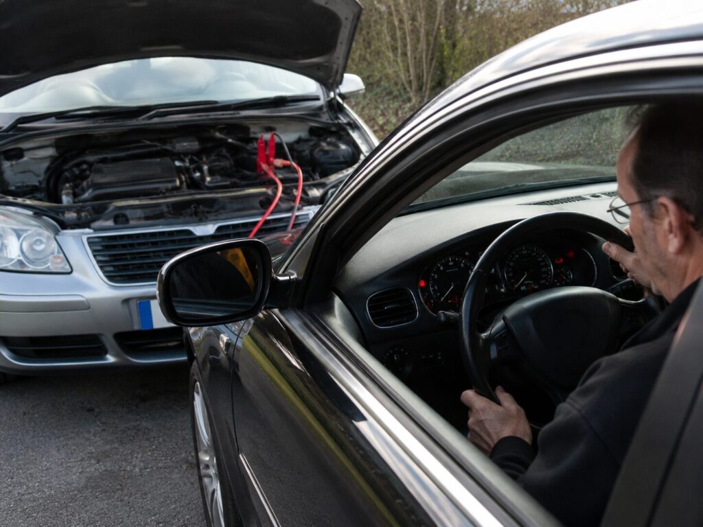 How to Jump Start A Car 17 Driver seated inside a car attempting to start the engine while another vehicle with its bonnet open is connected via jump leads in front. The scene shows a vehicle battery jump start in progress, with the driver checking the dashboard after receiving power from a donor vehicle.