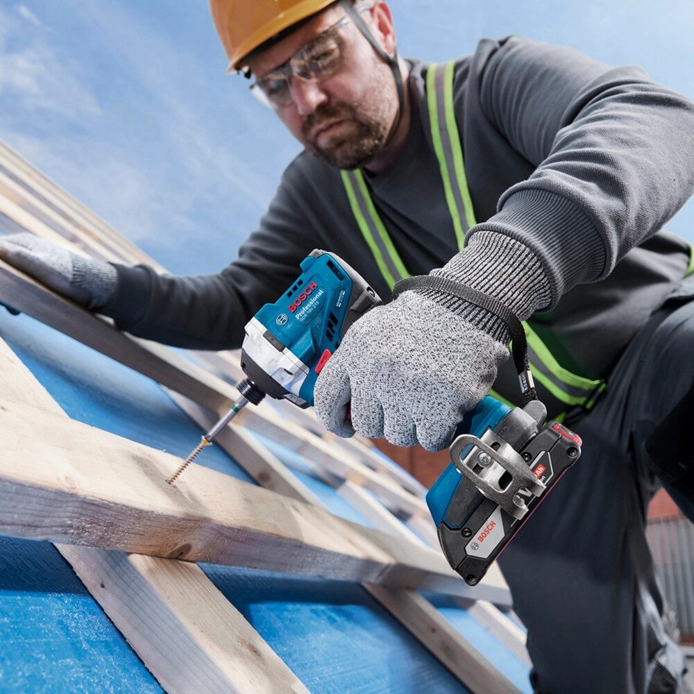 Construction worker using a Bosch cordless impact driver to drive a long timber screw into roof battens during roofing installation while wearing safety gloves, eye protection and a hard hat.